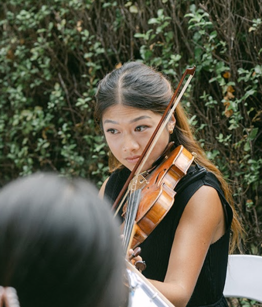 Young woman playing a violin outdoors with greenery in the background.