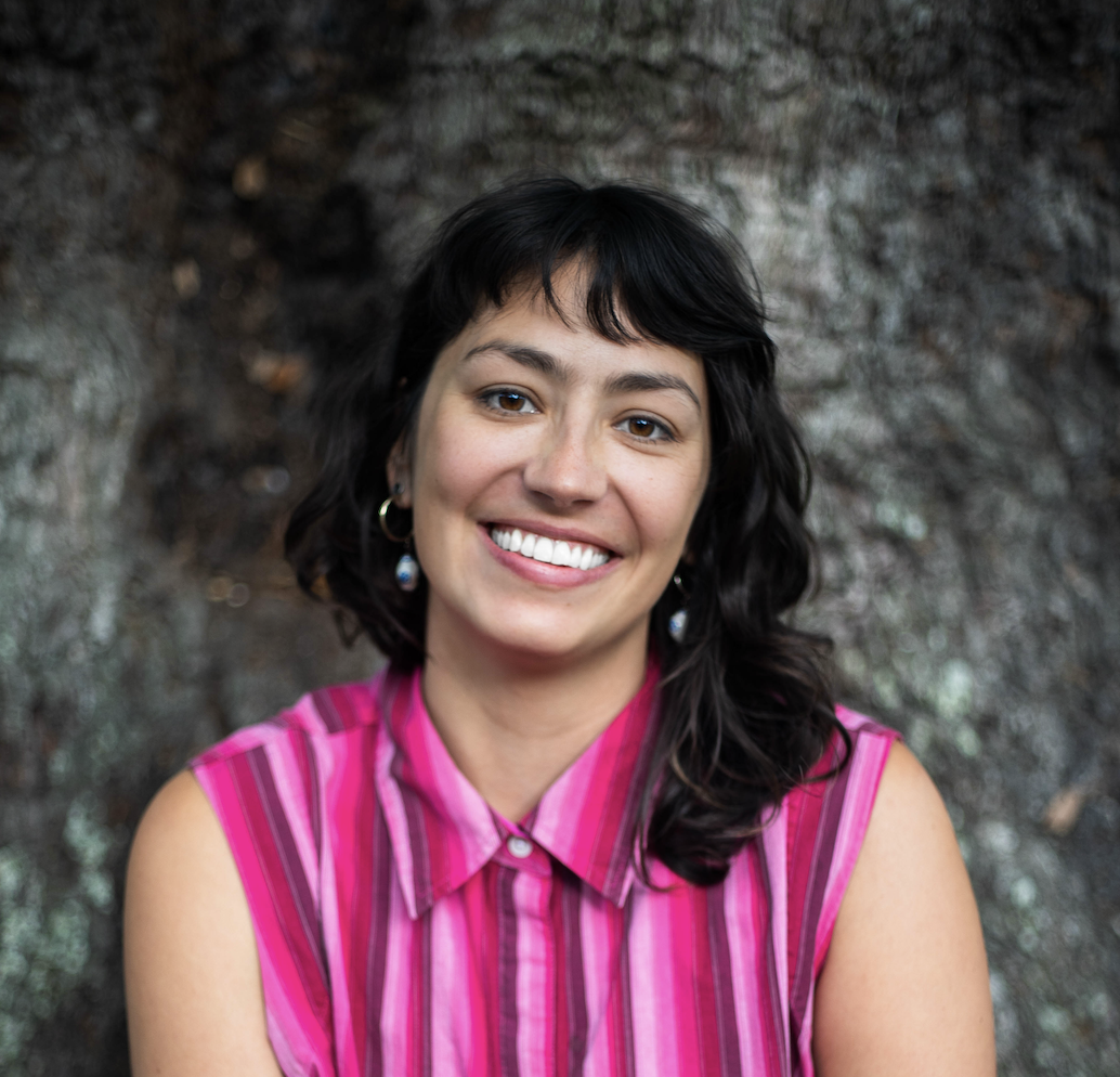 Emily Madeira, LMSW, a person in sleeveless white shirt smiling, standing in front of a wall with red shutters and greenery.