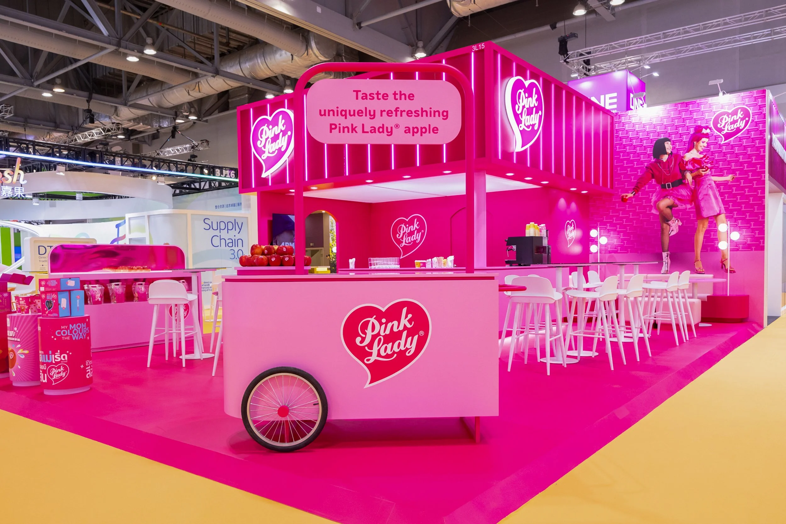 Pink Lady stand at Asia Fruit Logistica Exhibition, in Hong Kong, featuring a pink cart with the Pink Lady logo, a seating area with white chairs, a bar counter with apples.