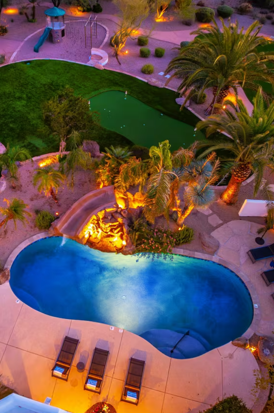 Night view of a luxurious outdoor pool area with a pool slide, palm trees, lawn, a small golf putting green, and lounge chairs, illuminated by soft lighting.