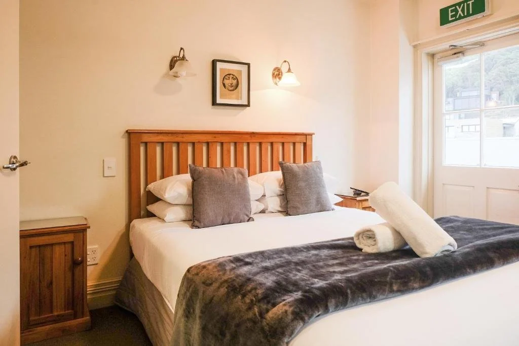 Hotel room with a large bed, green patterned pillows, a framed ocean-themed picture on the wall, a corner window with green leafy curtains, and a wooden dresser with a white electric kettle and coffee supplies.