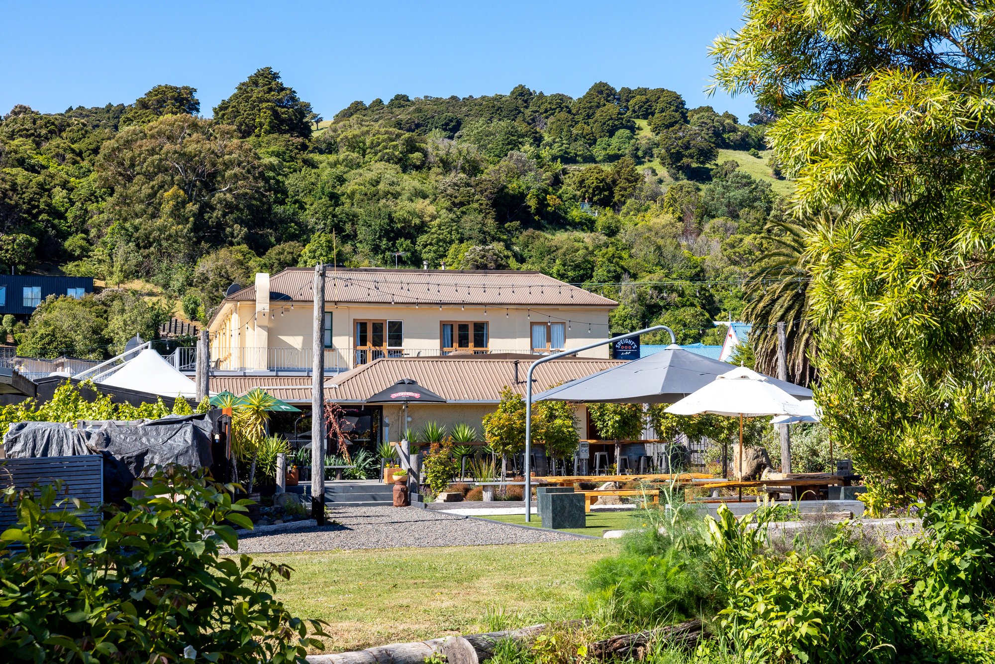 A scenic outdoor patio area with umbrellas, potted plants, a grassy lawn, and a building with a metal roof, set against lush green hills and trees under a clear blue sky.