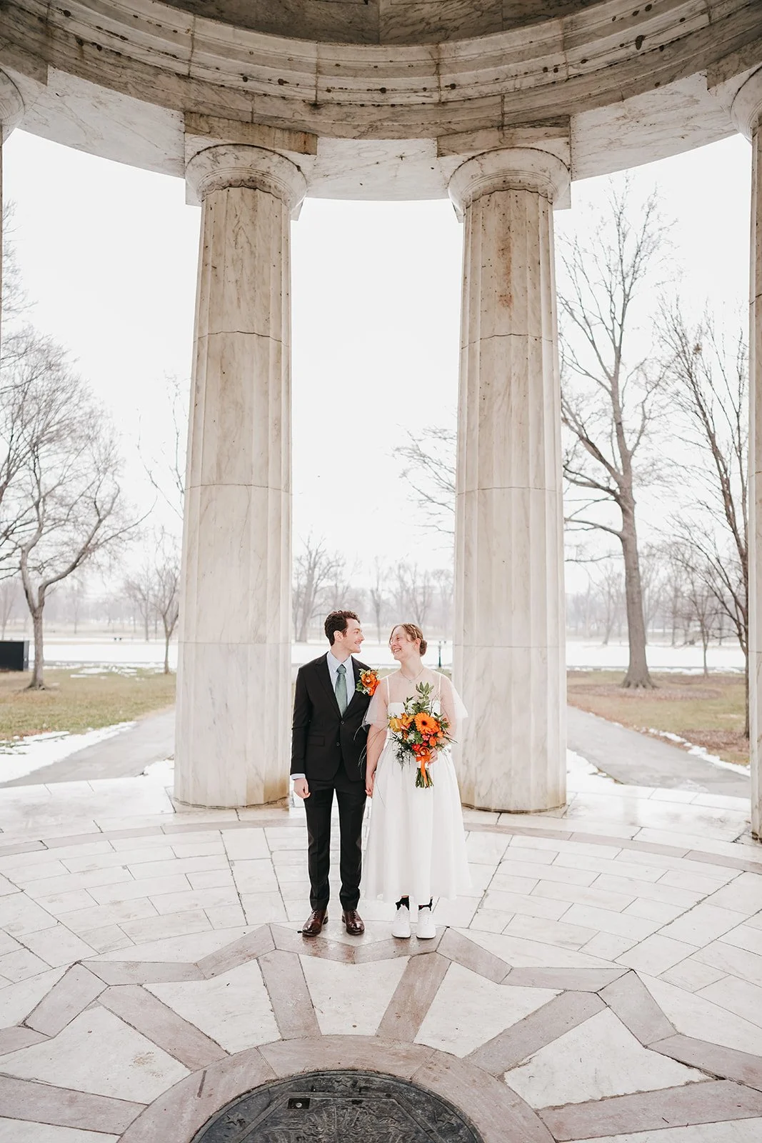 A couple in wedding attire standing under a large marble rotunda, holding hands and smiling at each other, with trees and snow in the background.