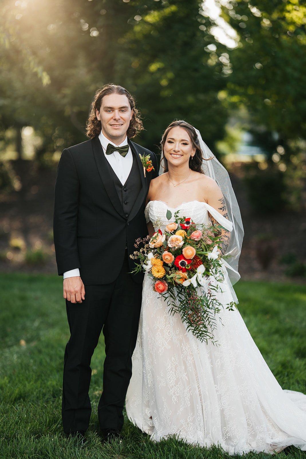 A newlywed couple standing outdoors on a grassy area with trees in the background. The groom is wearing a black tuxedo with a bow tie, and the bride is in a white wedding dress holding a colorful bouquet of flowers.
