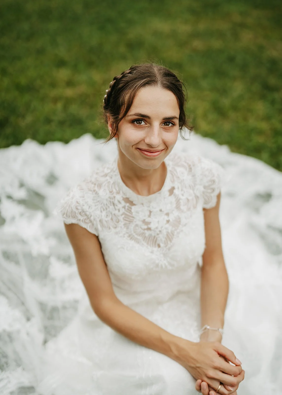 A young woman in a white lace wedding dress sitting outdoors on the grass, smiling at the camera.