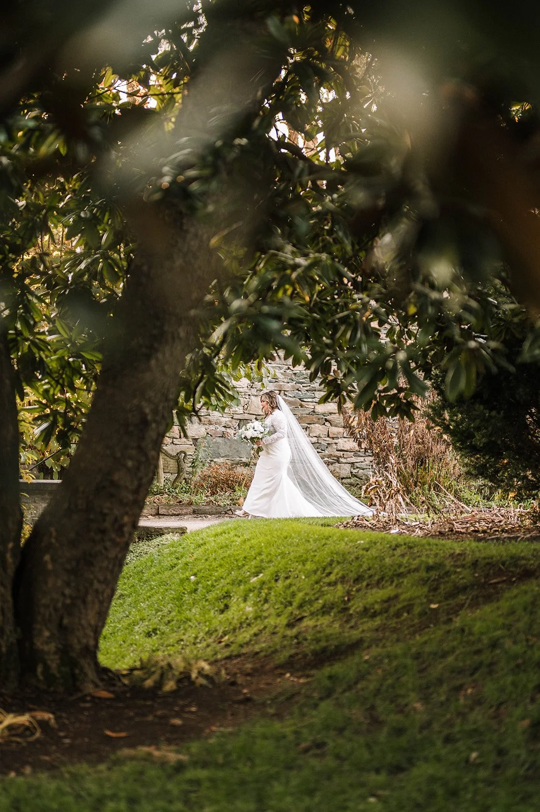 A bride in a white wedding gown and veil walking through a garden, holding a bouquet, with a stone wall in the background.