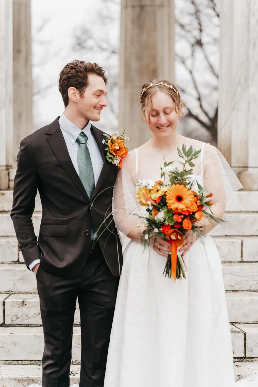 A bride and groom standing on steps outdoors, the bride holding a bright orange bouquet, both smiling softly, in front of a structure with columns and a leafless tree in the background.