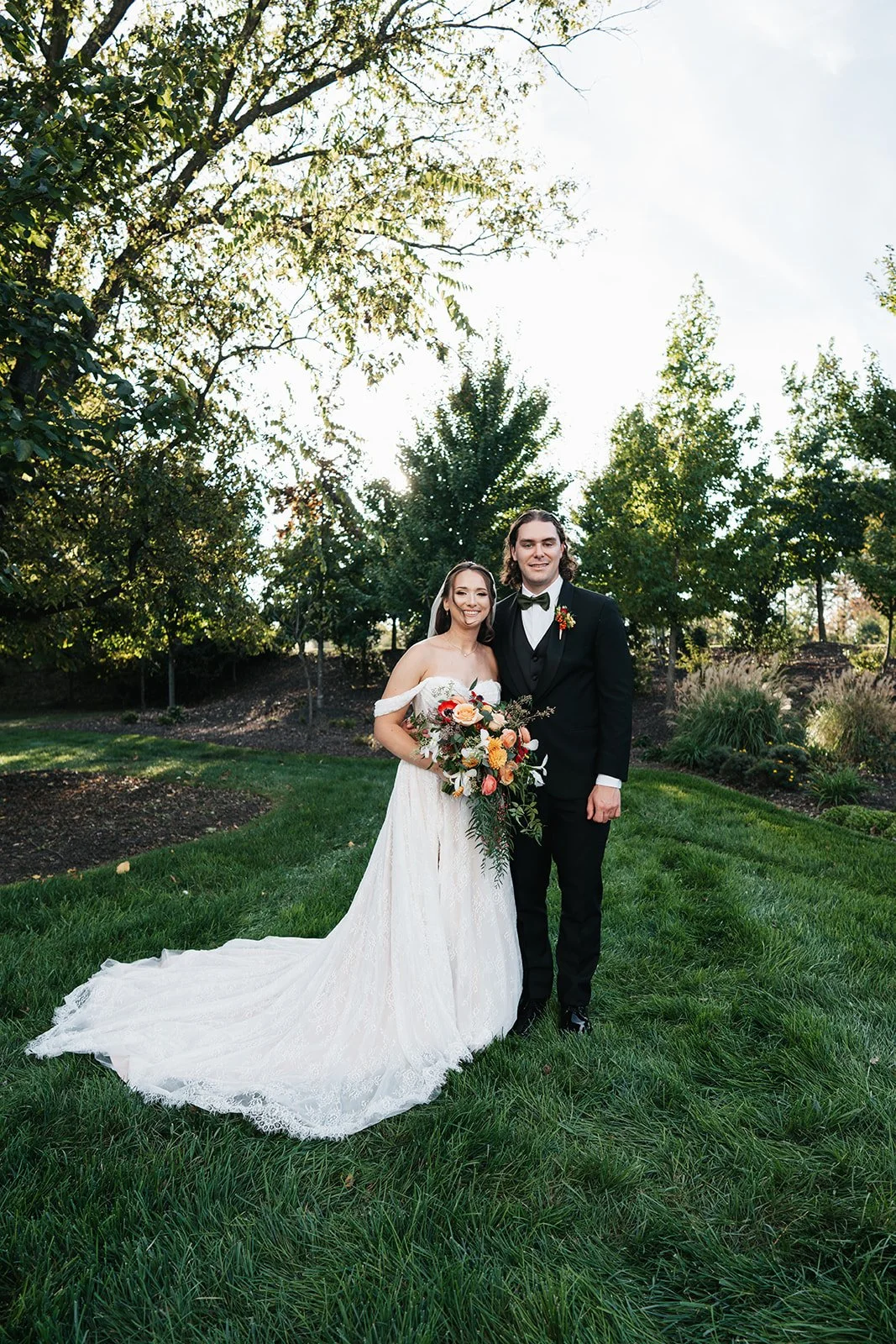 A bride and groom standing together on a grassy lawn surrounded by trees, with the bride holding a bouquet of flowers and both smiling at the camera.
