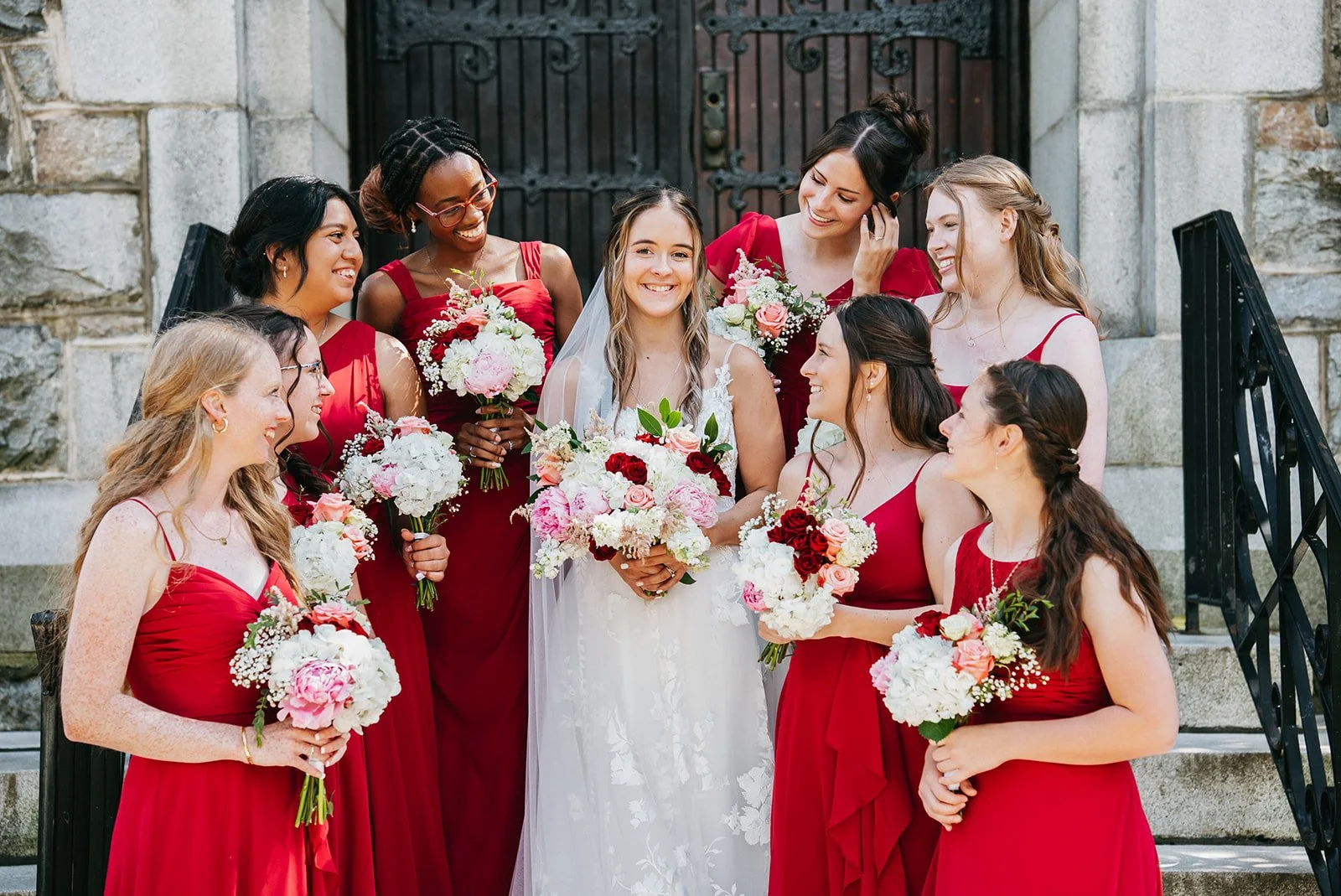 A bride in a white wedding dress holding a bouquet, surrounded by nine bridesmaids in red dresses holding bouquets, standing on stone steps in front of a black iron door.
