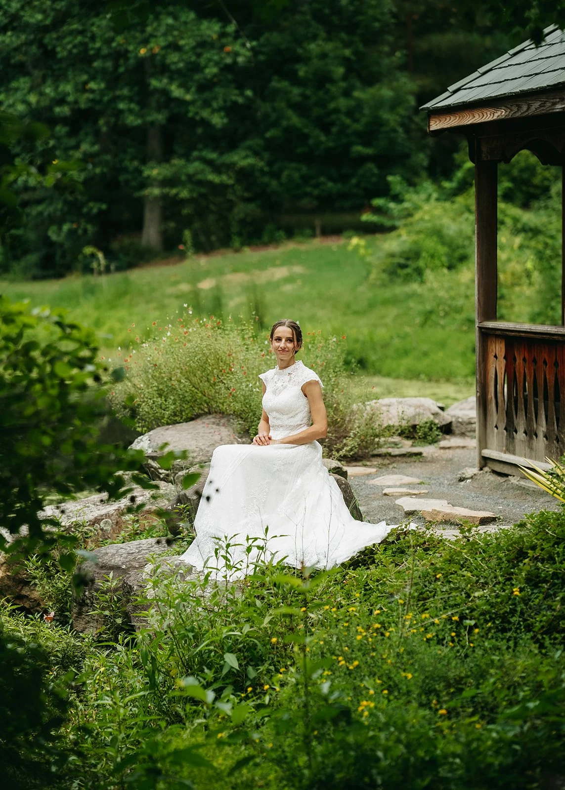 A woman in a white wedding dress sitting on rocks near a garden gazebo, surrounded by lush greenery.