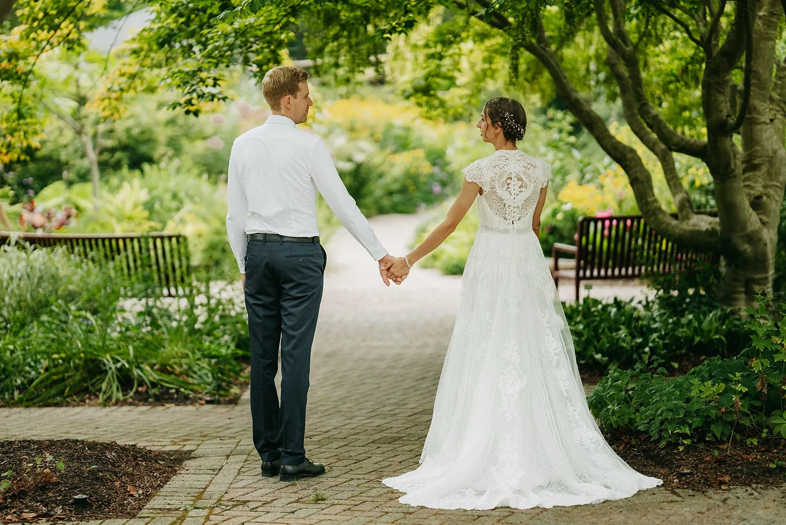 A bride in a white lace wedding dress and a groom in a white shirt and dark trousers holding hands in a lush garden.
