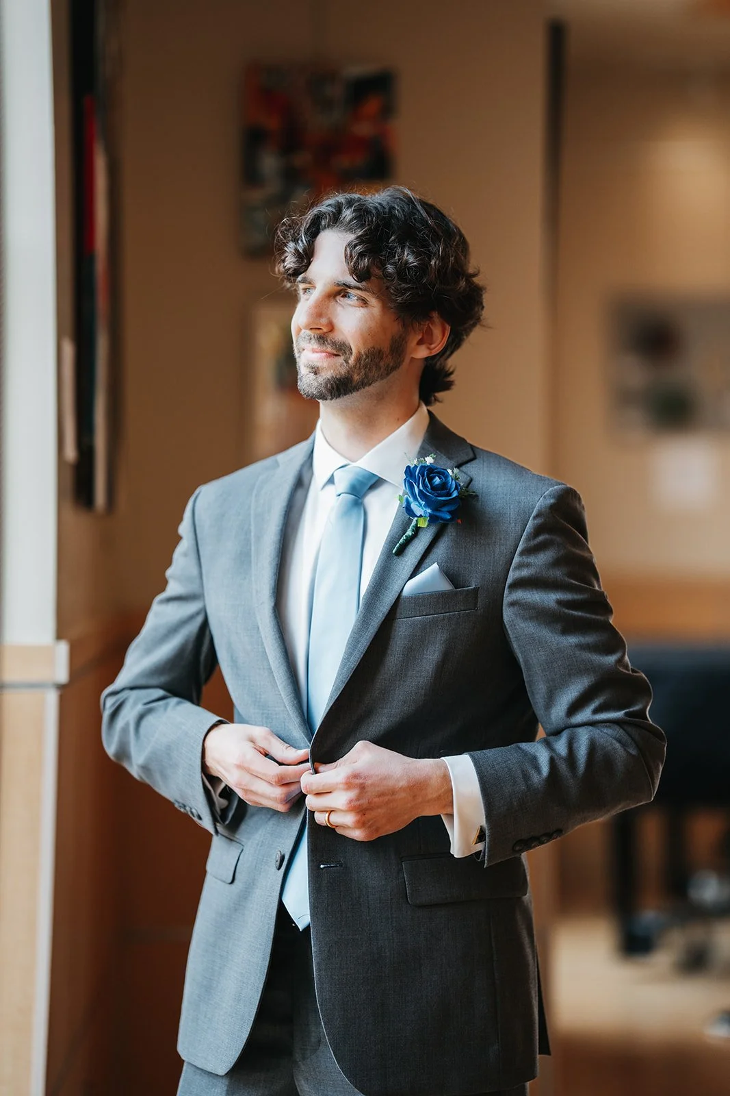 A man in a gray suit with a light blue tie and a blue rose boutonniere, standing indoors near a window with natural light, adjusting his jacket.