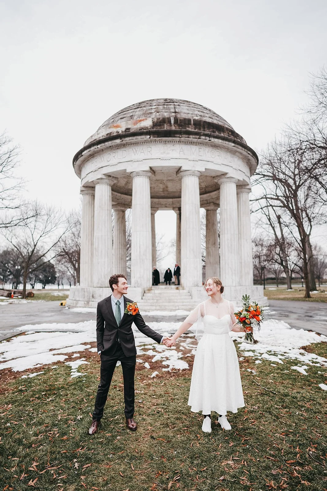 A bride and groom holding hands and smiling in front of a historic memorial with a domed roof and columns, on a cloudy day with snow patches on the ground.