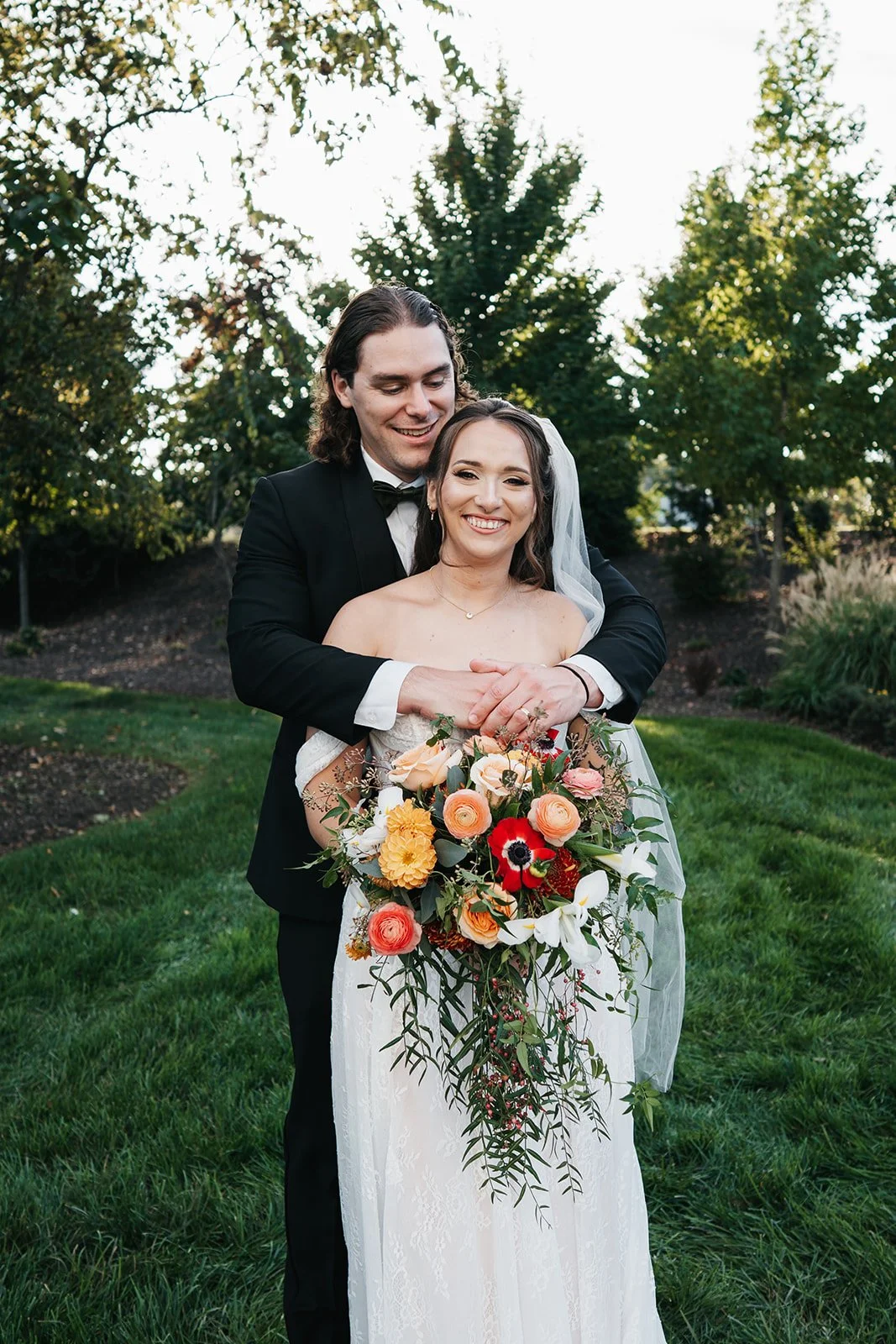 A newlywed couple stands outdoors on a grassy lawn, with the groom hugging the bride from behind, both smiling happily. The bride holds a large, colorful bouquet of flowers. In the background, there are trees and shrubbery.