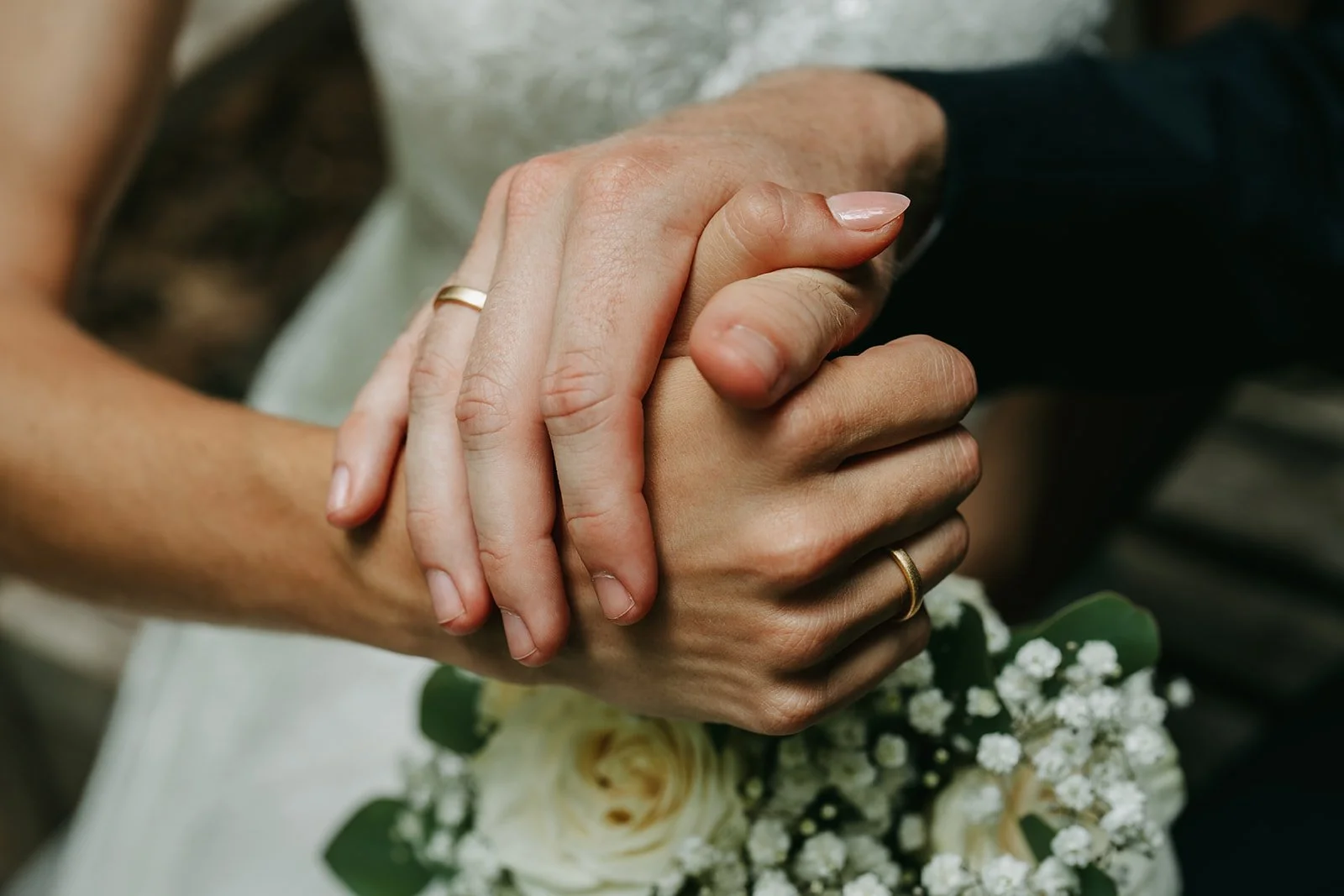 Close-up of a couple holding hands during a wedding, with wedding rings on their fingers and a bouquet of white flowers in the background.
