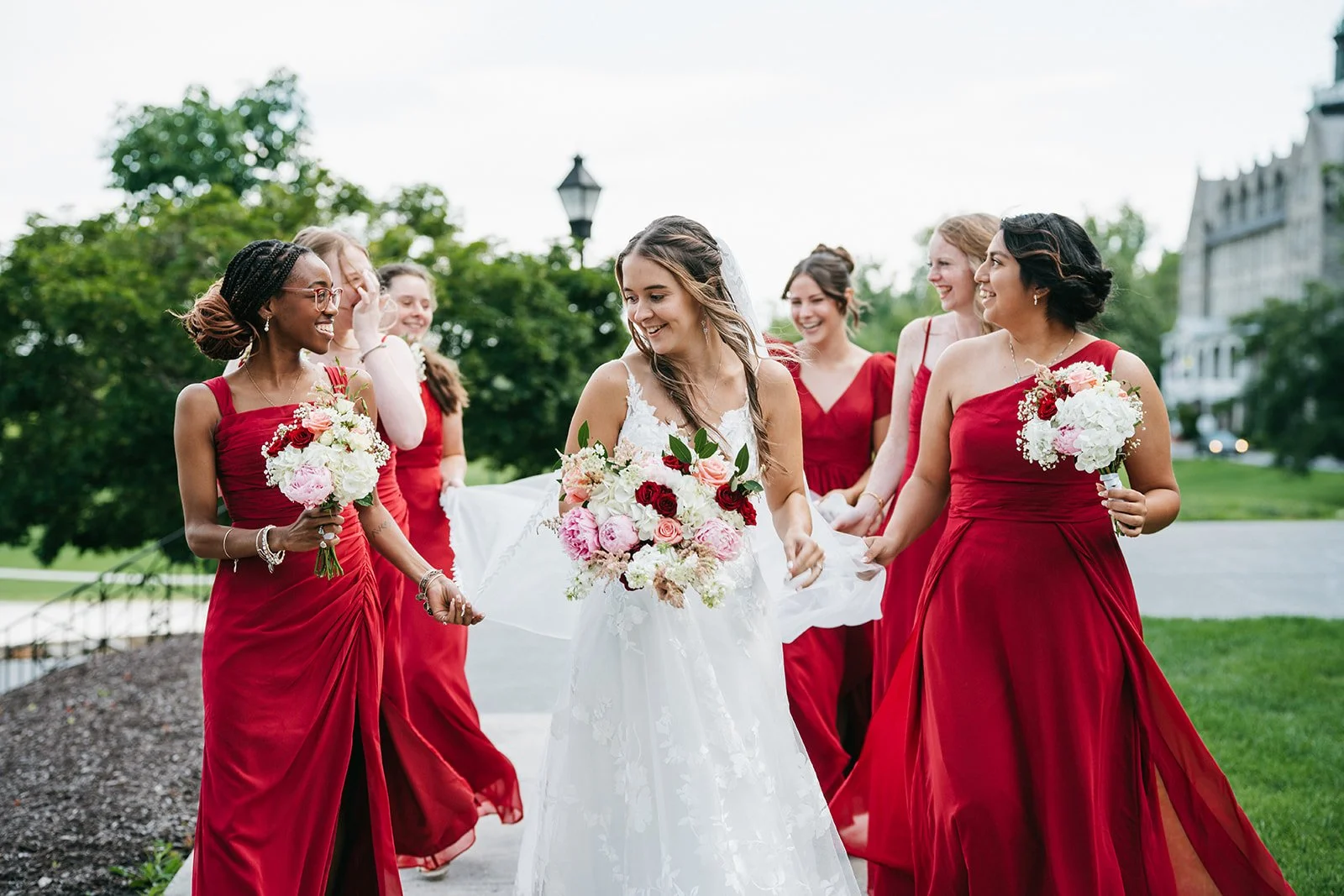 A bride in a white wedding dress walking with her bridesmaids dressed in red dresses, all holding floral bouquets in a park setting.