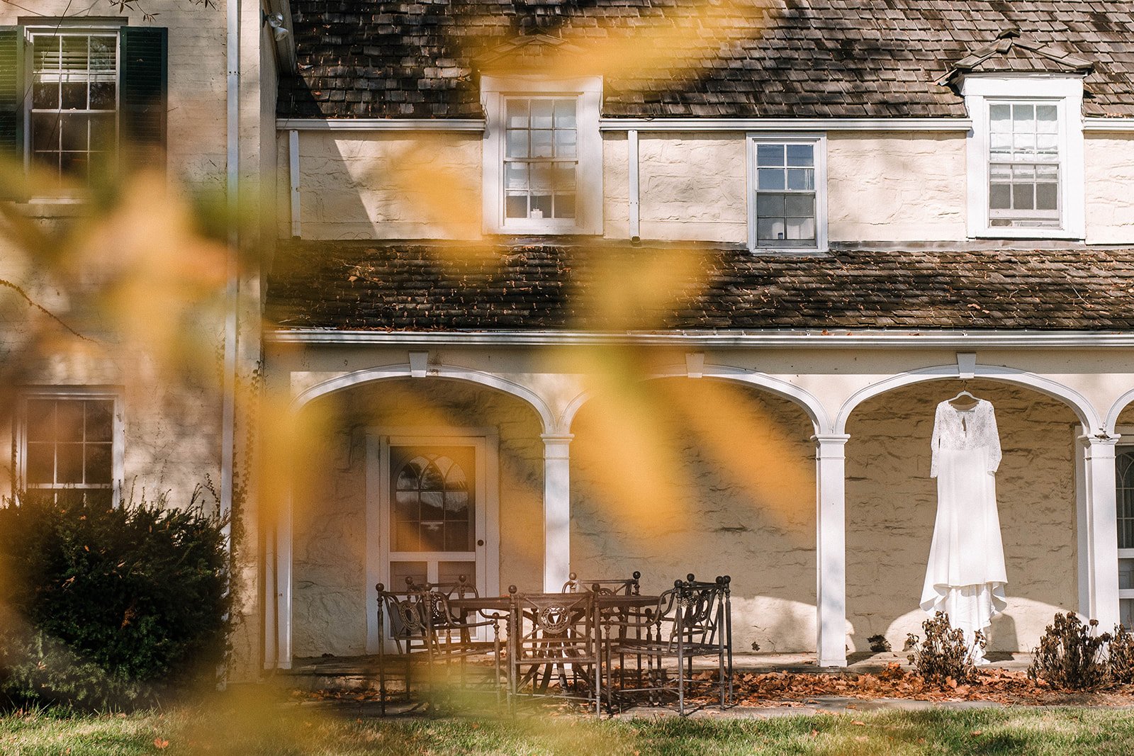 A white wedding dress hanging outside on a porch of an old house with arched columns, brown roof shingles, and multiple windows, with autumn leaves in the foreground.