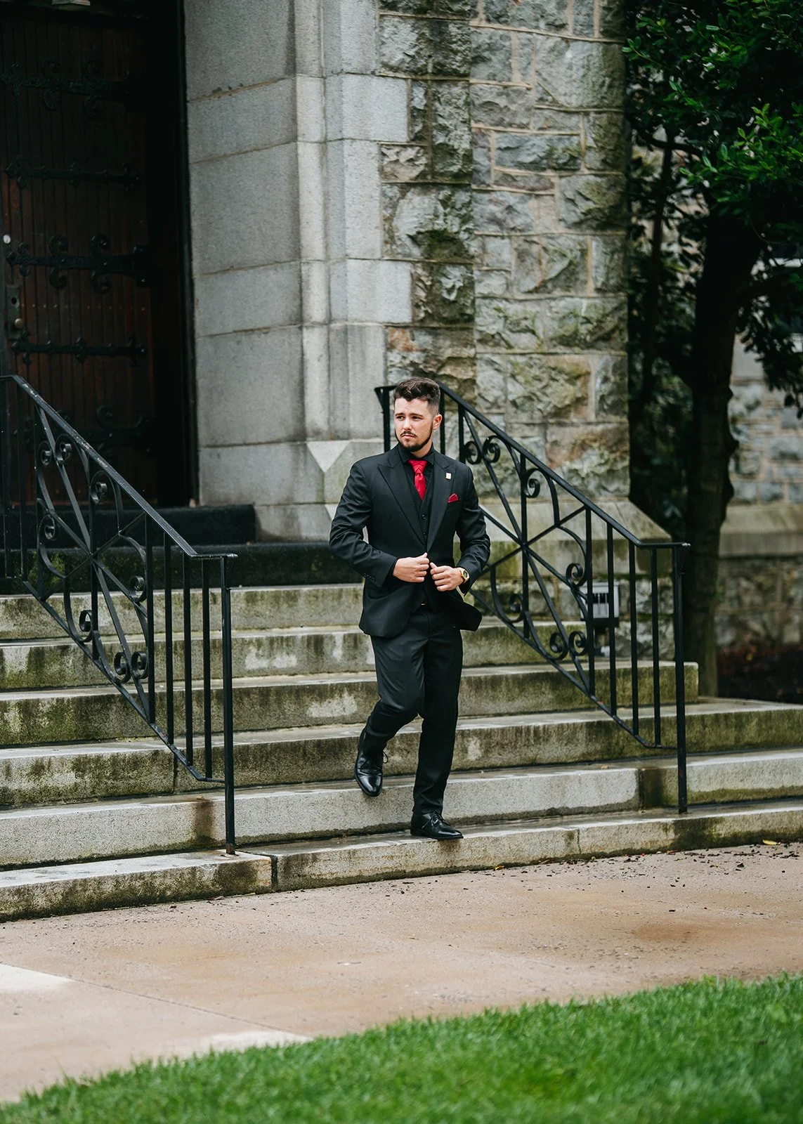 A man in a black suit with a red tie and pocket square walking down stone steps outside a building with stone and brick wall, black iron stairs railing, and green trees in the background.