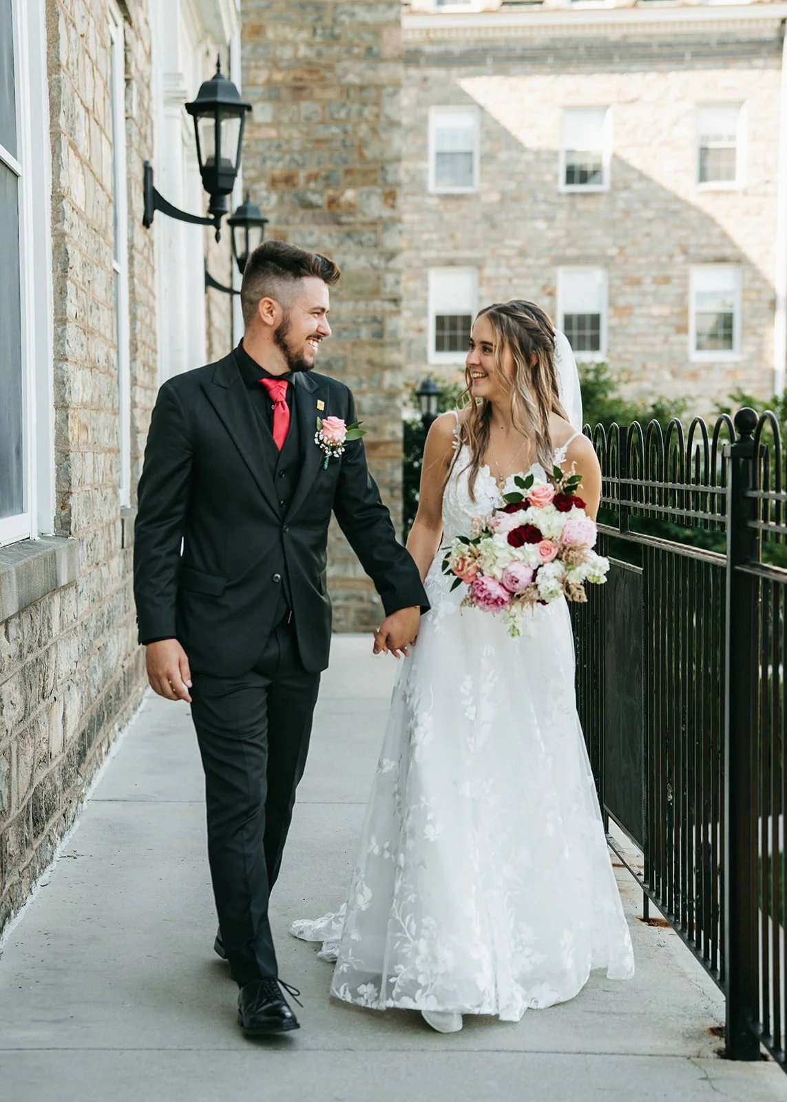 A bride and groom walking hand in hand outdoors on their wedding day. The groom is wearing a black suit with a red tie and boutonniere, and the bride is in a white wedding dress holding a colorful bouquet of flowers. They are smiling at each other ne