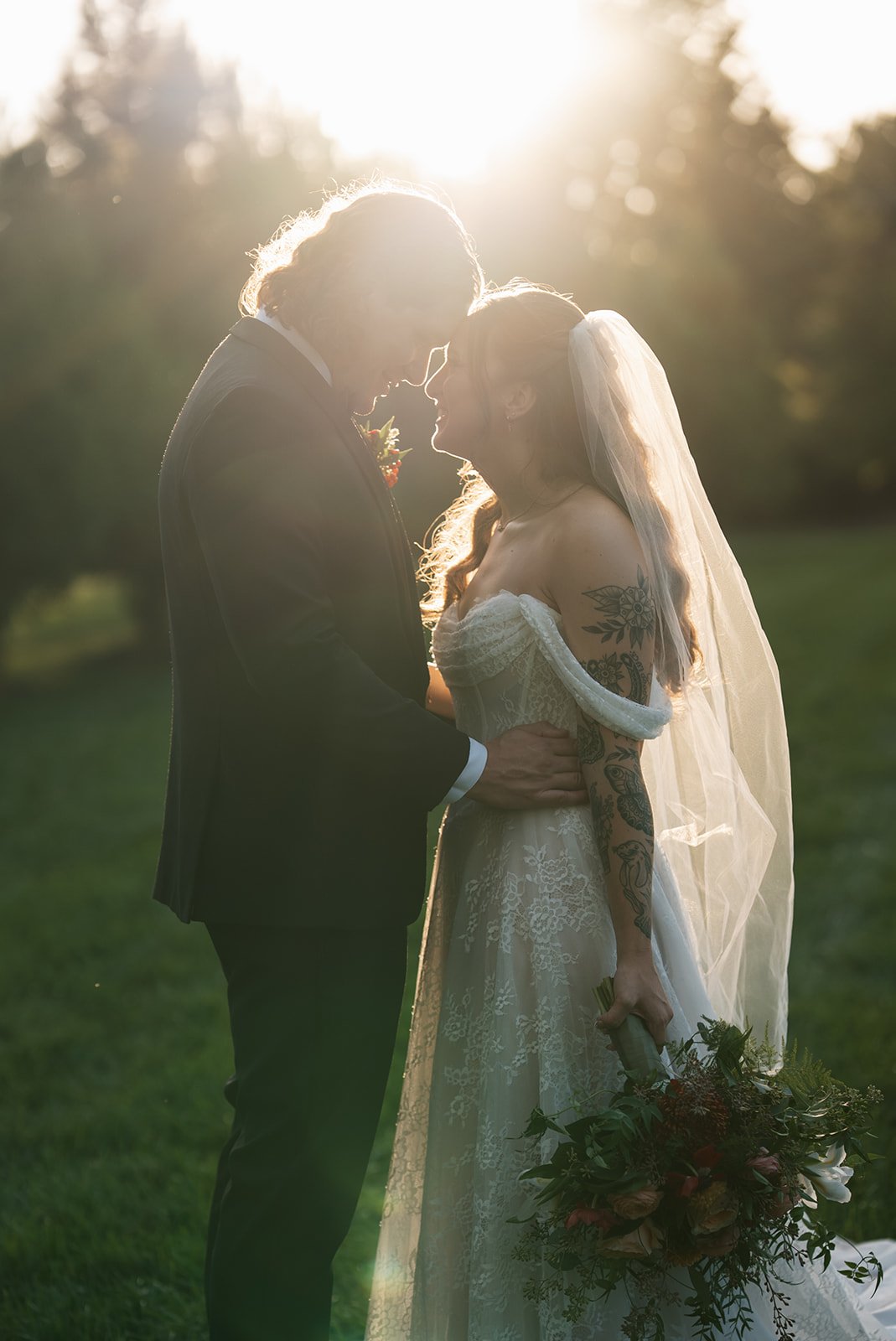 A newlywed couple in wedding attire stands close together outdoors at sunset, holding hands and smiling, with the bride holding a bouquet of flowers.