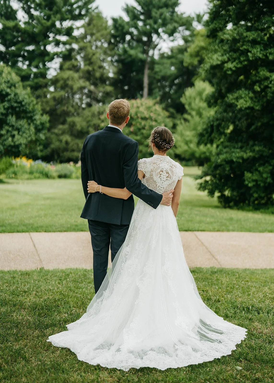 A bride and groom standing arm-in-arm outdoors, facing away from the camera, surrounded by trees and greenery, with the bride in a white lace wedding gown and the groom in a black suit.