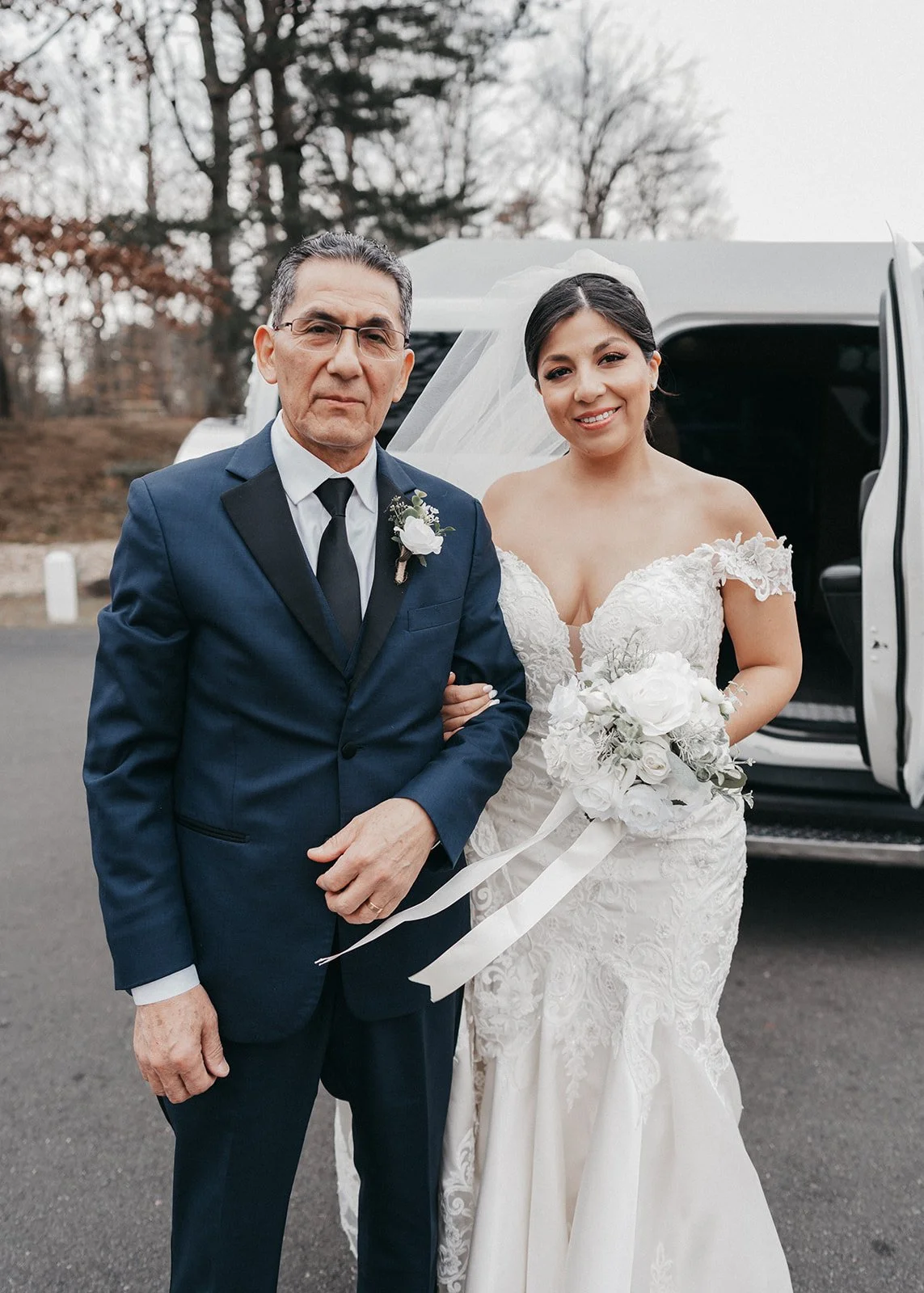 Bride in a white wedding dress holding a flower bouquet standing next to a man in a blue suit and black tie in front of a white vehicle.