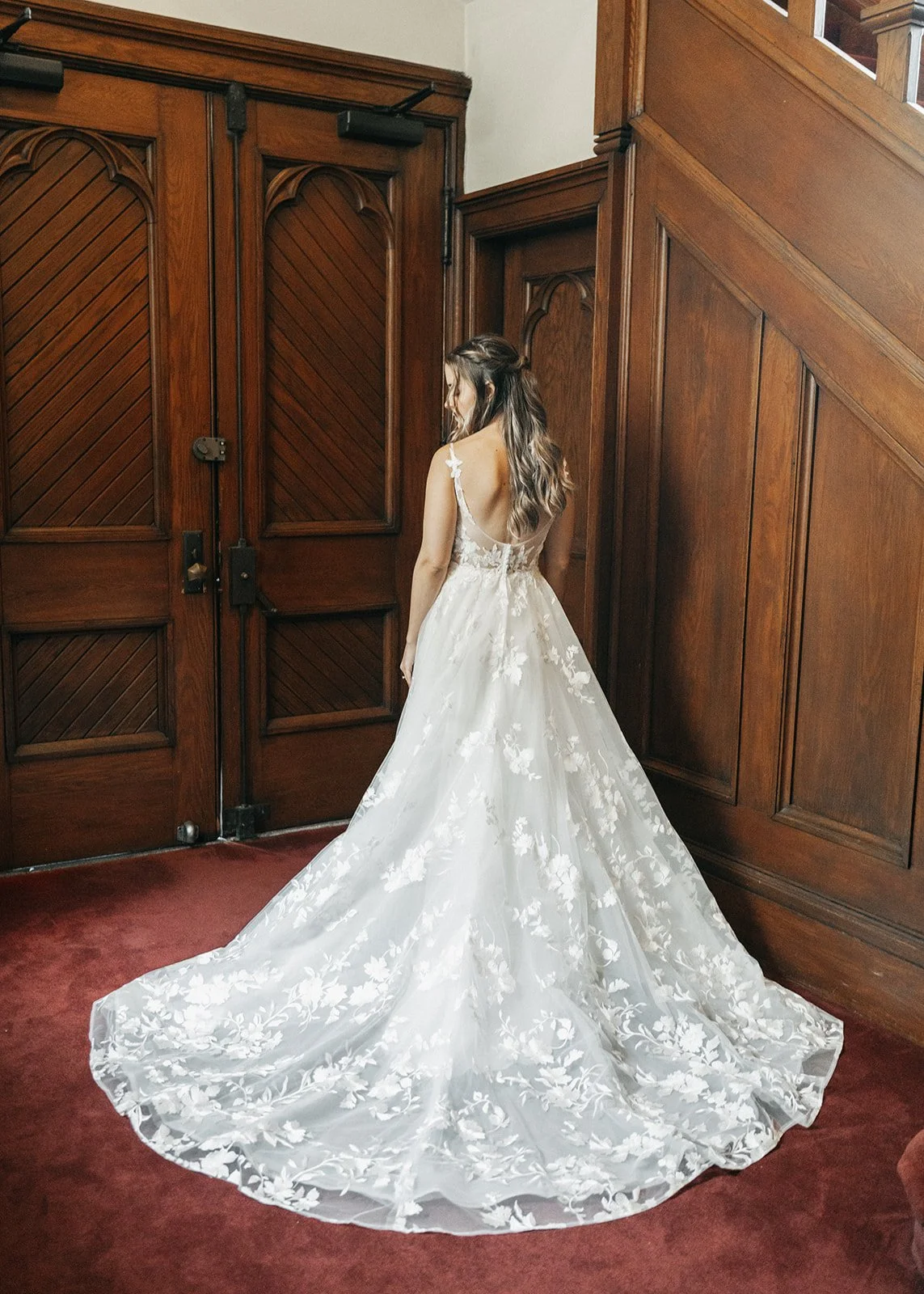 A woman in a white wedding dress with floral embroidery stands with her back to the camera, facing a wooden door and wall, in a room with a red carpeted floor.