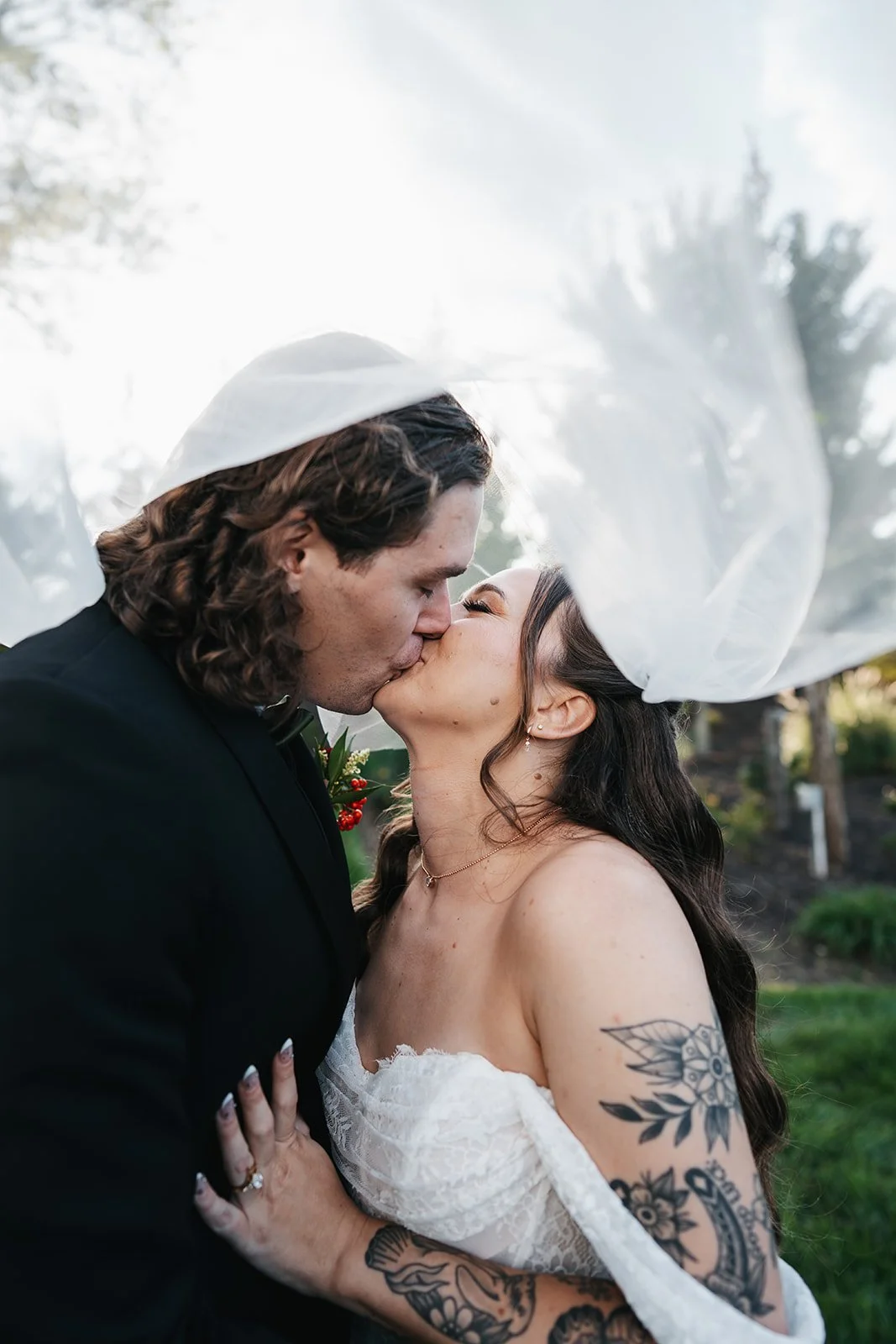 A bride and groom kiss outdoors, with the bride wearing a veil and a strapless wedding dress, and it appears to be a wedding or special occasion.