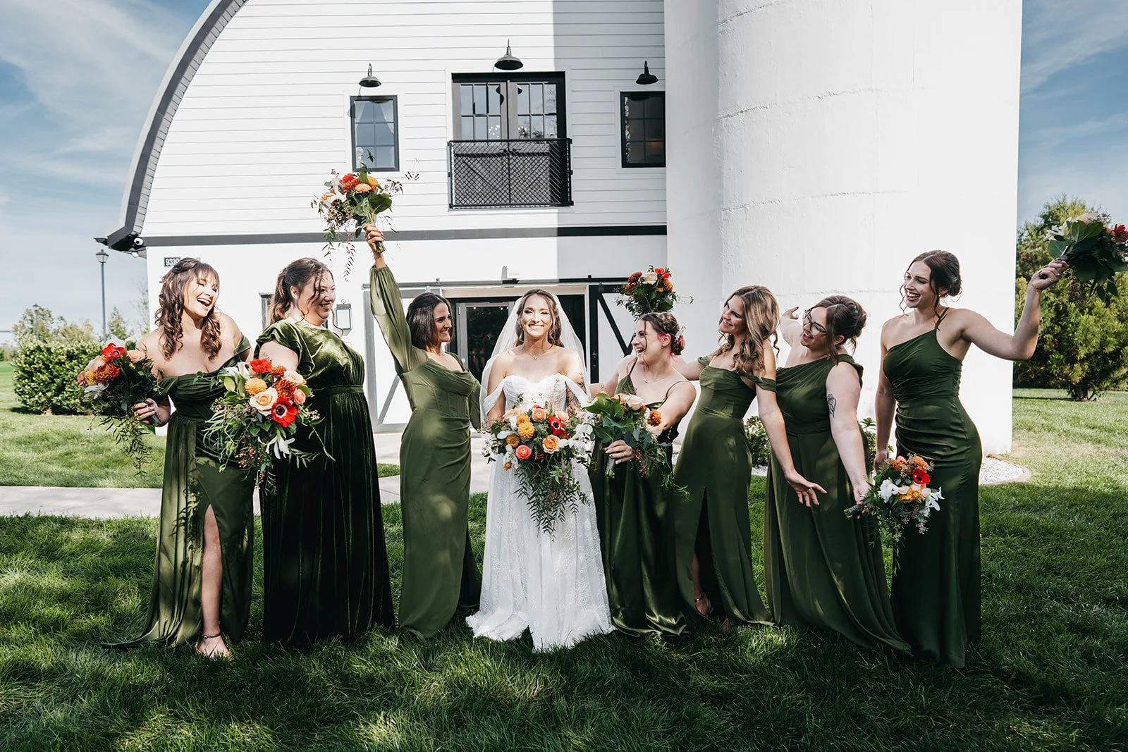 A group of nine women, including a bride in a white wedding dress with a veil, and eight bridesmaids in dark green dresses, are standing outside on a grassy lawn in front of a white building, celebrating with bouquets of colorful flowers. The bride i