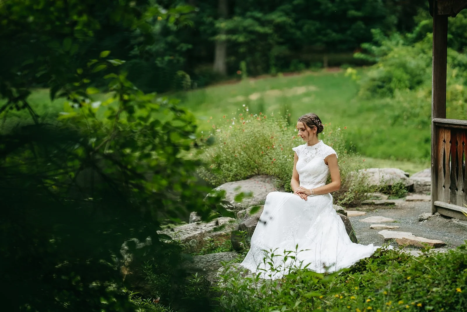 A bride in a white lace wedding dress sitting on rocks in a lush green garden.