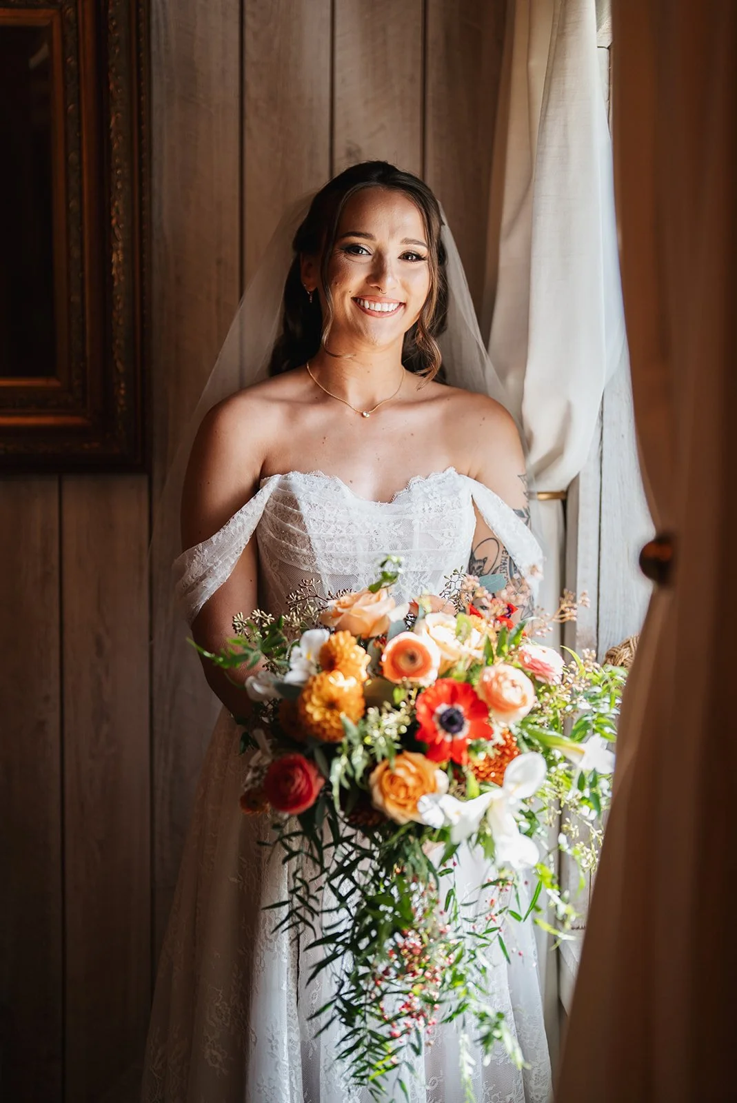 A smiling bride in a wedding dress holding a large colorful bouquet, standing by a window with wooden walls and curtains.