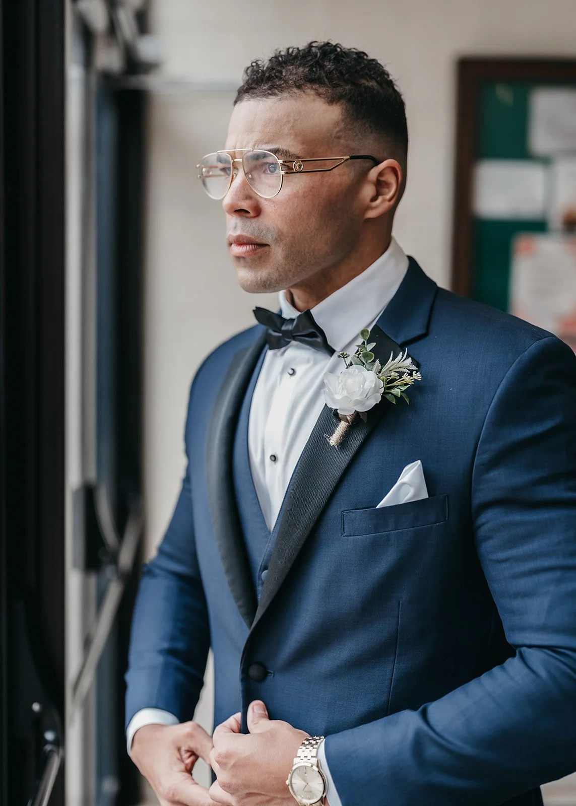 A man dressed in a navy blue suit and white shirt, wearing a black bow tie and glasses, looking out a window. He has a white boutonniere pinned to his lapel and a white pocket square, with a silver watch on his left wrist.