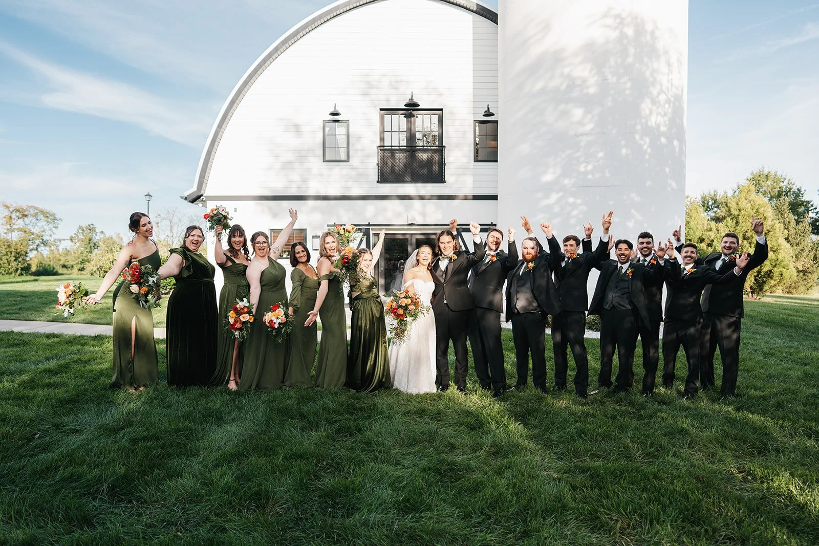 A group of bridesmaids and groomsmen celebrating outside a white barn on a sunny day. The women are wearing green dresses and holding bouquets, while the men are in black suits with boutonnières, and they are all smiling and raising their arms.