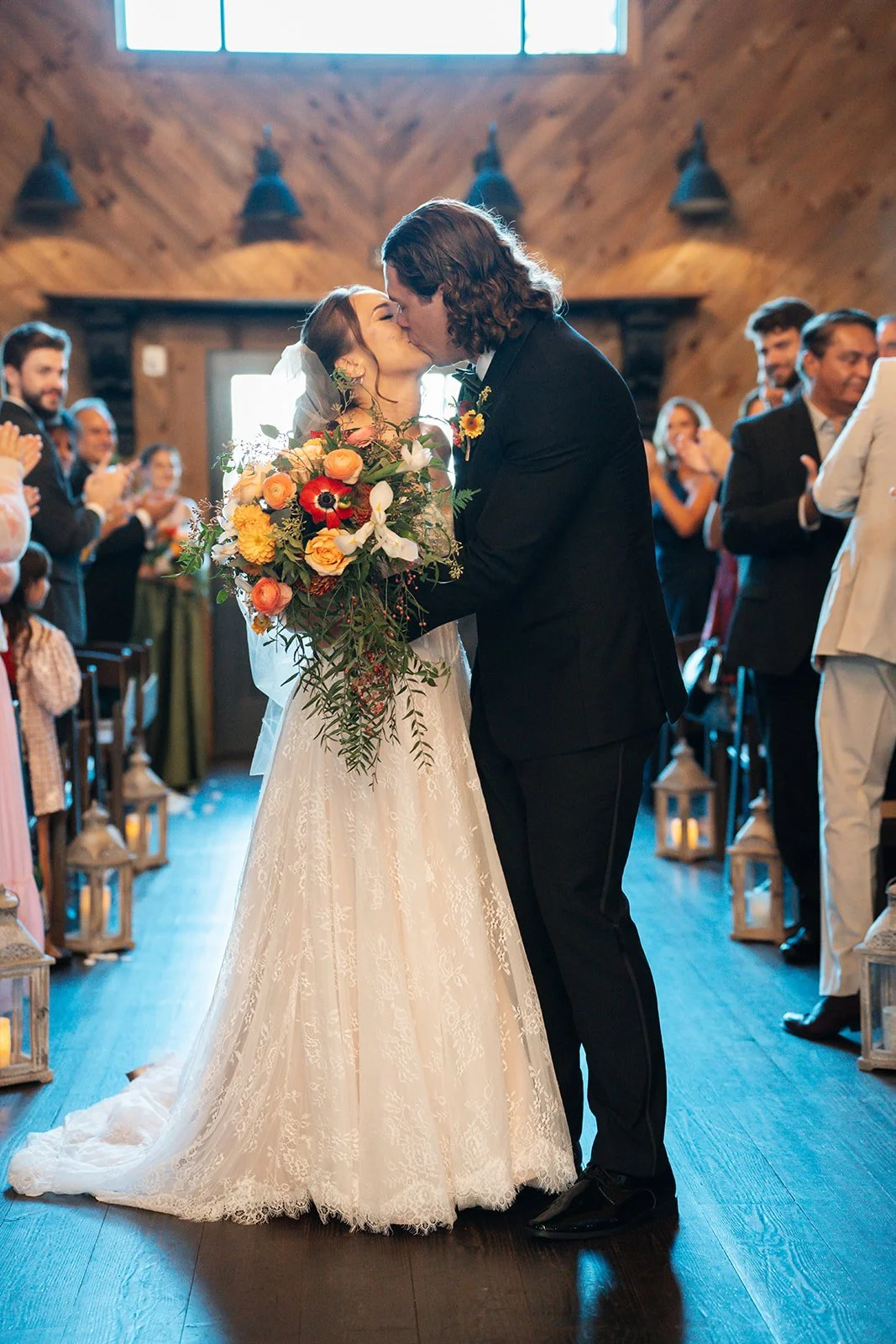 A bride and groom sharing a kiss during their wedding ceremony in a rustic indoor venue with wooden walls, surrounded by friends and family.