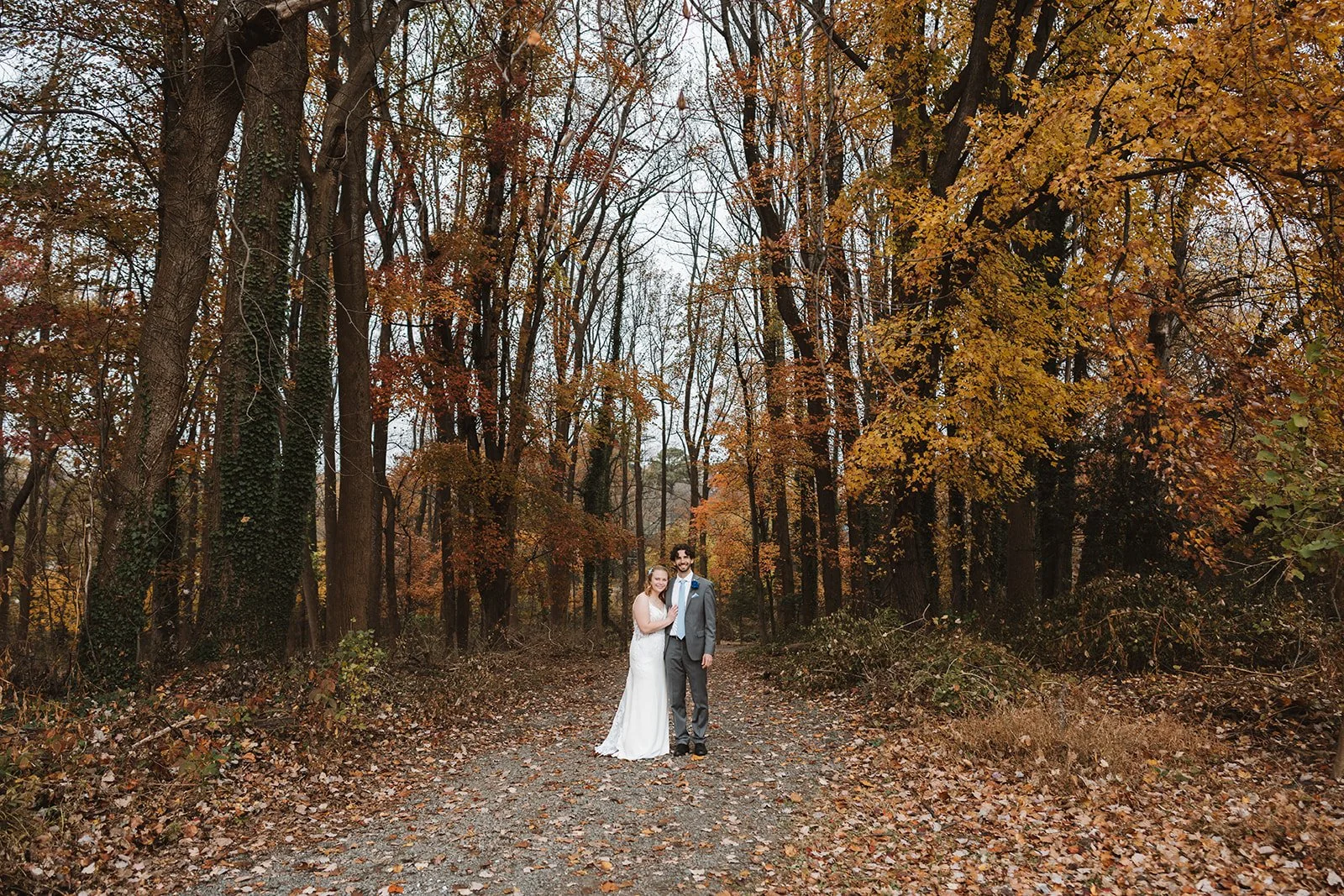 A bride and groom standing on a forest path surrounded by autumn trees with orange and yellow leaves.