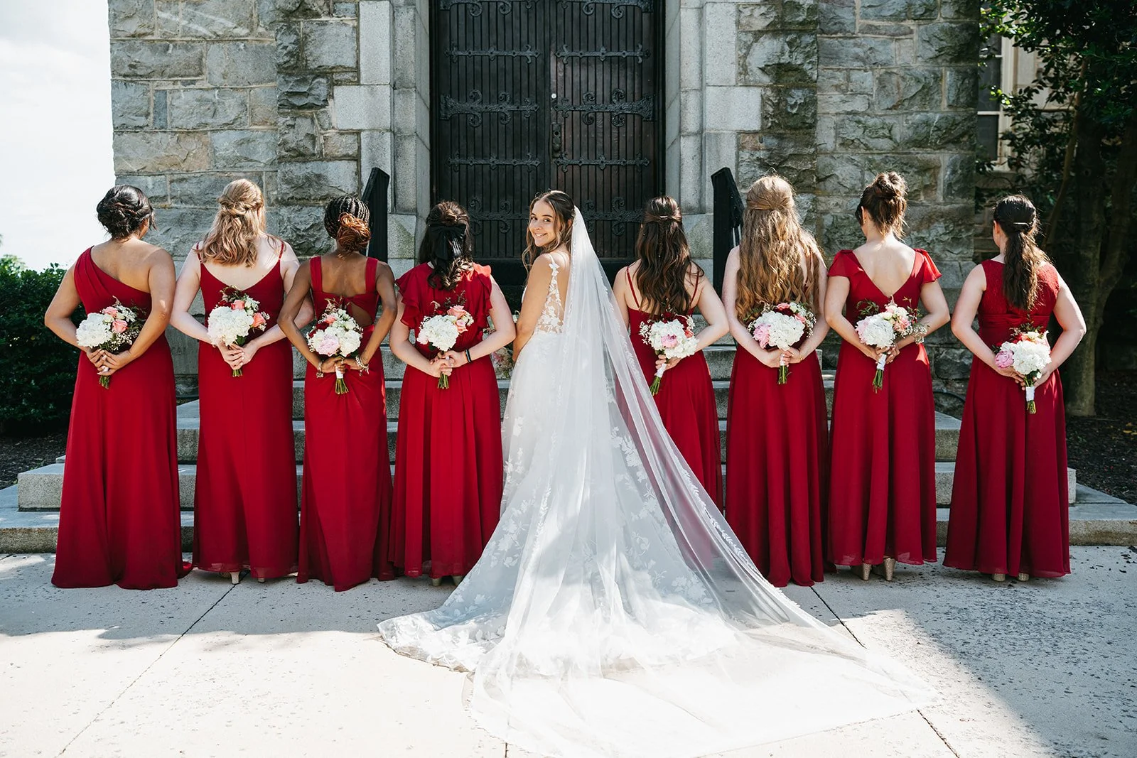 A bride in a white wedding gown with a long train standing with her back to the camera, facing a line of bridesmaids in red dresses. The bridesmaids hold bouquets of pink and white flowers and are positioned on steps in front of a stone building with