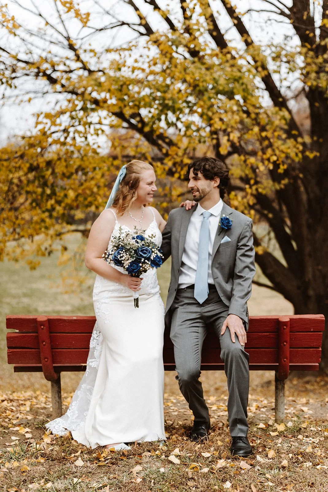 A bride and groom sitting on a park bench, smiling at each other, with autumn leaves and a large tree with yellow leaves in the background. The bride is holding a bouquet of blue and white flowers, and the groom is dressed in a gray suit with a blue 