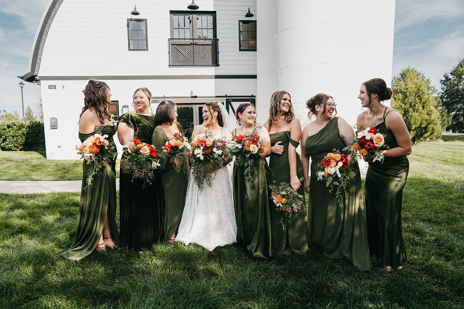 A group of eight women, including a bride, in formal dresses with dark green and black hues, holding colorful bouquets, standing on a lawn outside a white building with a curved roof. They are smiling and looking at each other.