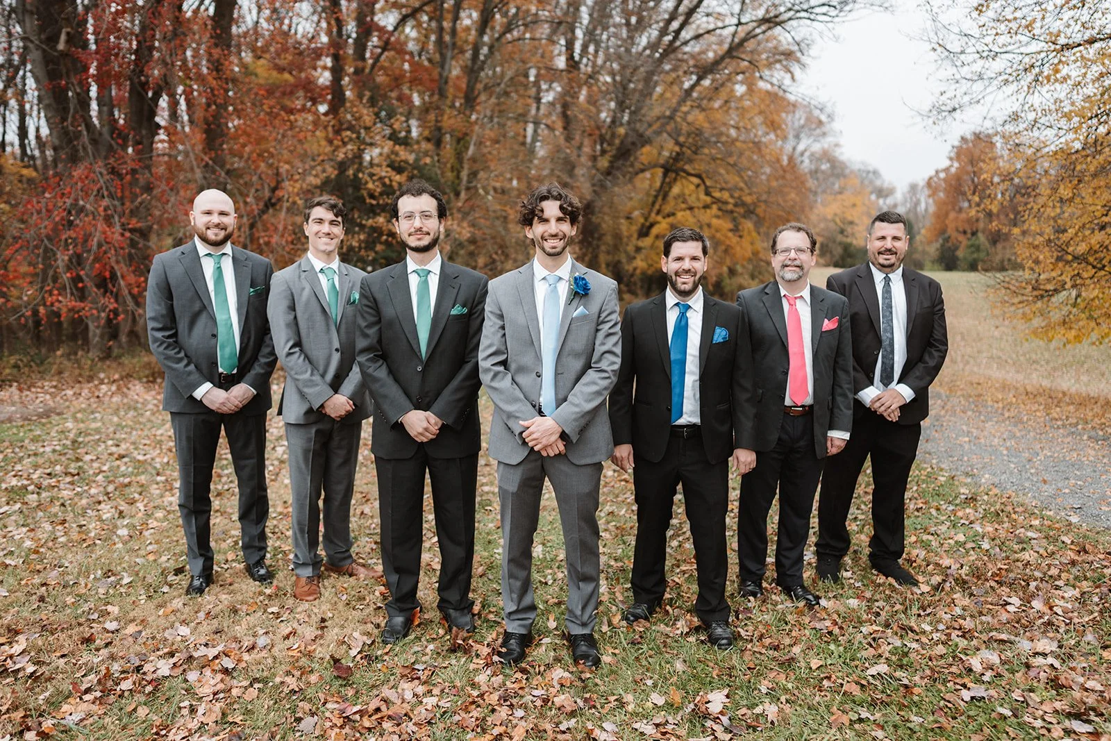A group of seven men in suits standing outdoors on a leaf-covered ground during autumn, smiling for a photo.