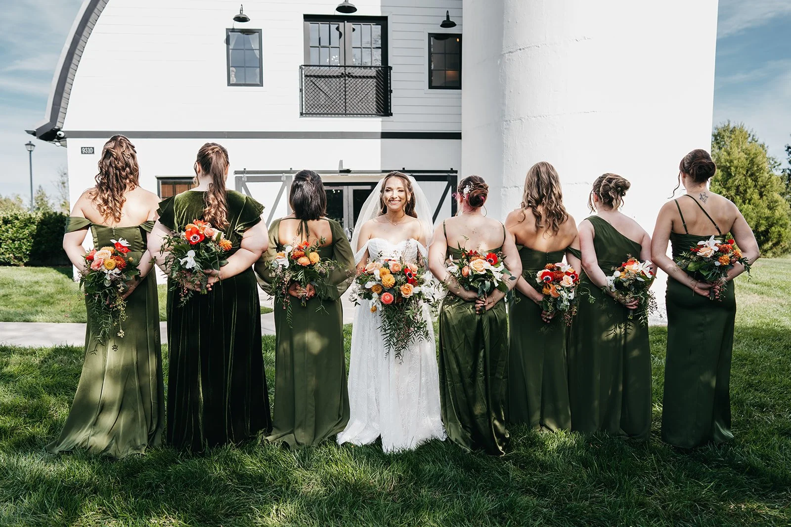 A bride in a white wedding dress and seven bridesmaids in matching dark green dresses and floral bouquets, standing outdoors in front of a modern white building with a rounded roof.