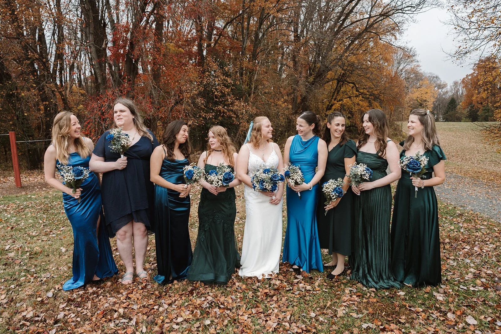 Wedding party of ten women standing outdoors among fall foliage, some in blue and some in green dresses, holding bouquets of blue and white flowers, smiling and talking.
