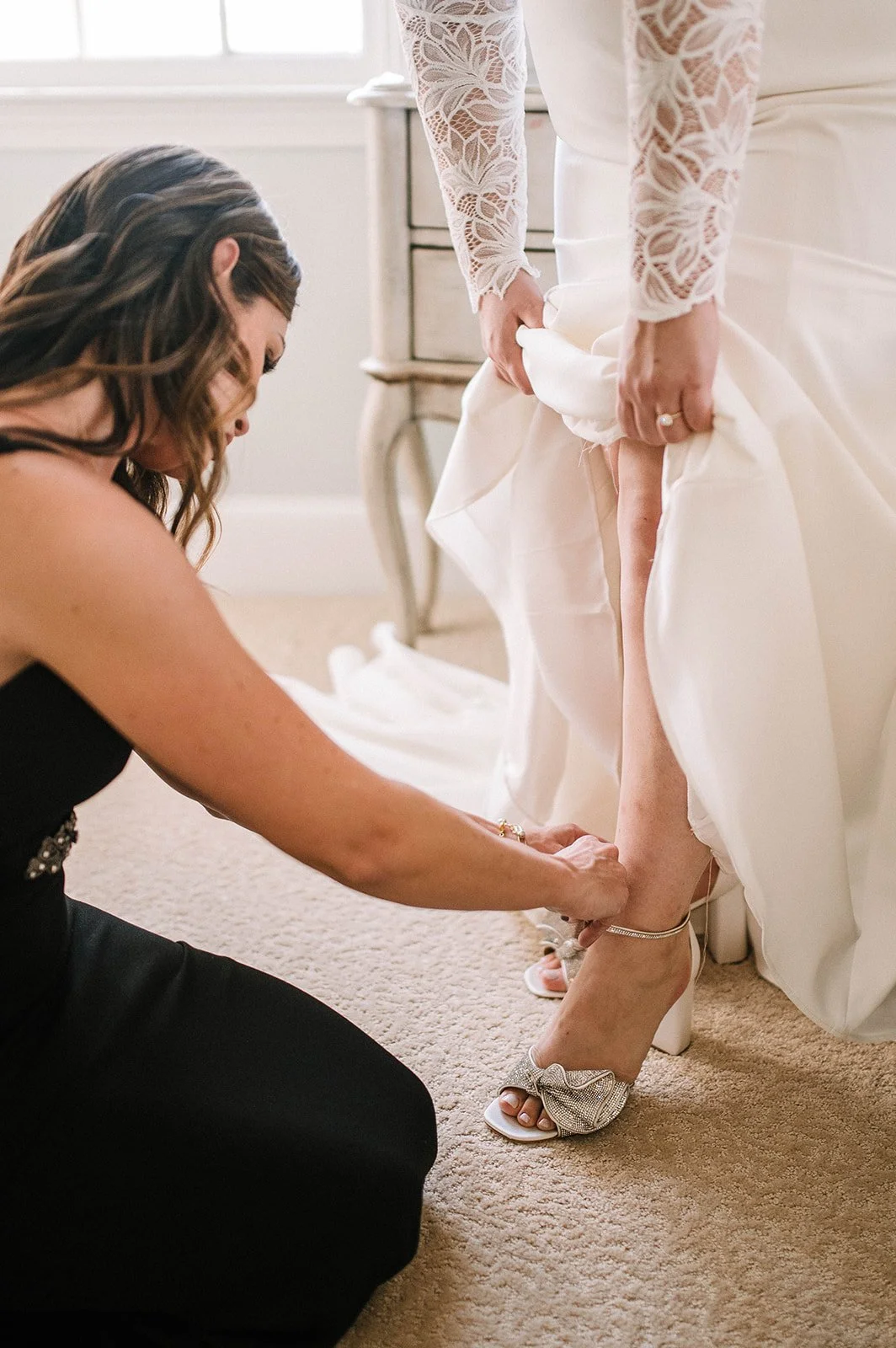 A woman in a black dress helping another woman in a white wedding gown put on a high-heeled shoe with a bow.