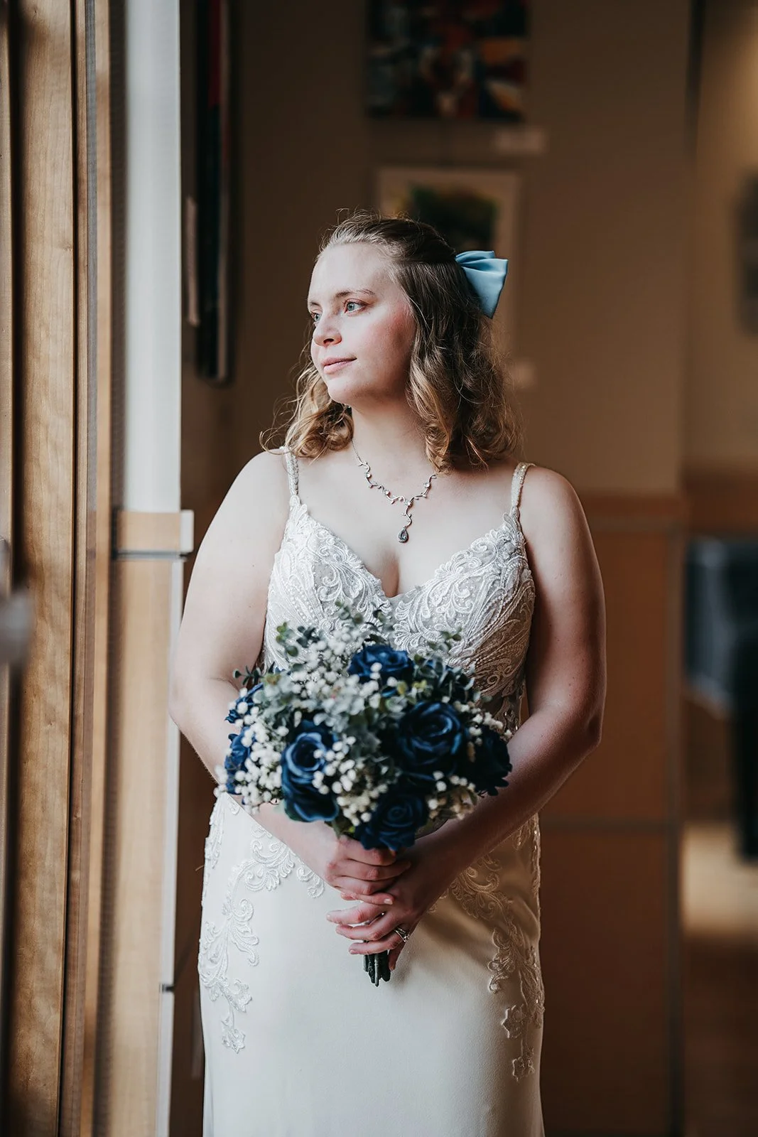 A bride in a white wedding dress with lace details, holding a bouquet of blue roses and white flowers, standing indoors near a window with a contemplative expression.