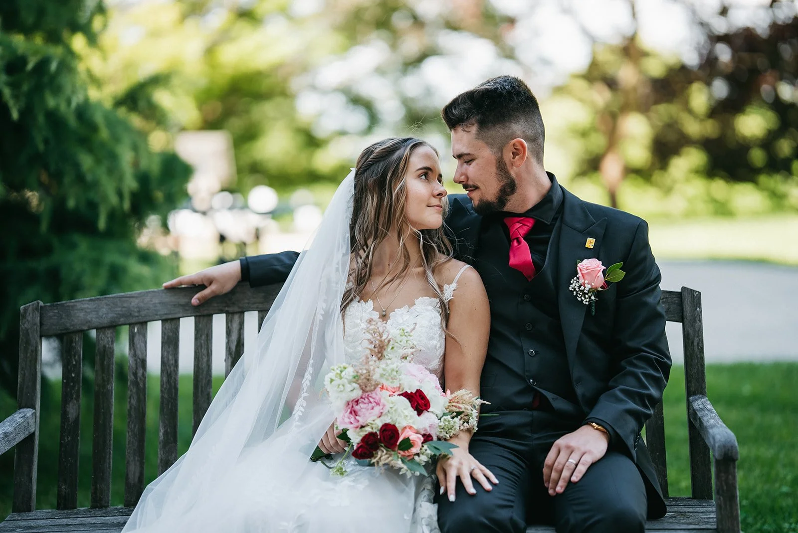 A bride and groom sitting closely on a wooden bench outdoors, gazing into each other's eyes, with the bride holding a bouquet of flowers and greenery, surrounded by lush greenery and trees.