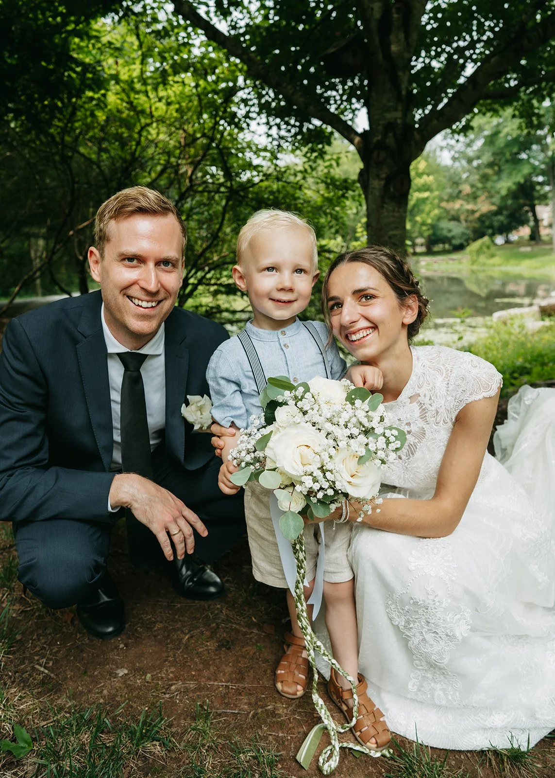 A happy family of three at a wedding outdoors, including a man in a dark suit, a woman in a white lace dress holding a bouquet, and a young boy in light clothing holding the bouquet.
