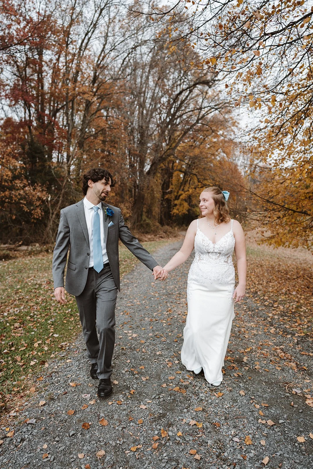 A bride and groom holding hands and walking outdoors on a fall day, surrounded by autumn trees with colorful leaves.
