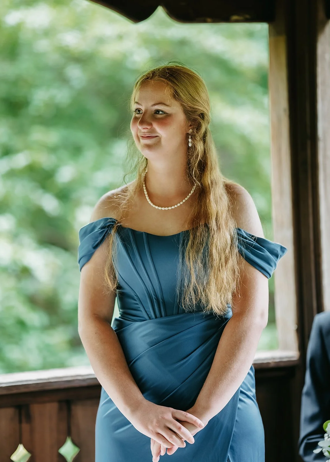 A woman with long, wavy blonde hair wearing a blue off-shoulder dress, pearl necklace, and earrings, standing indoors with a blurred green outdoor background visible through a window.