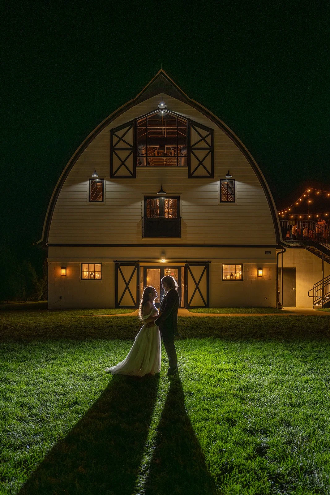 A bride and groom stand facing each other on a grassy lawn at night, illuminated from behind, with a large, barn-style building brightly lit in the background.