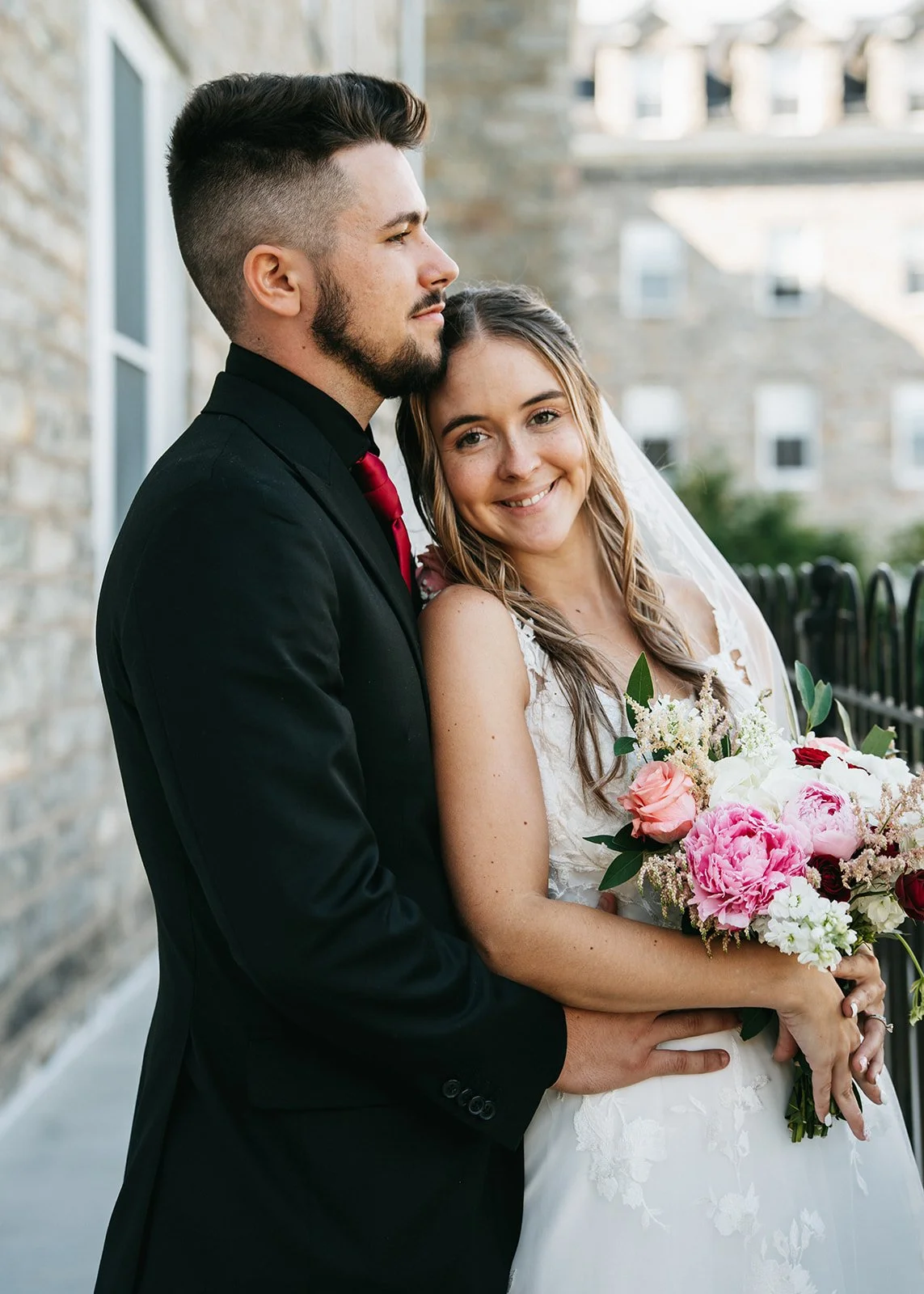 A newlywed couple stands outside, with the bride holding a bouquet of pink and white flowers. The groom wears a black suit with a red tie. They are smiling and close to each other, with the groom gently resting his arm around the bride.