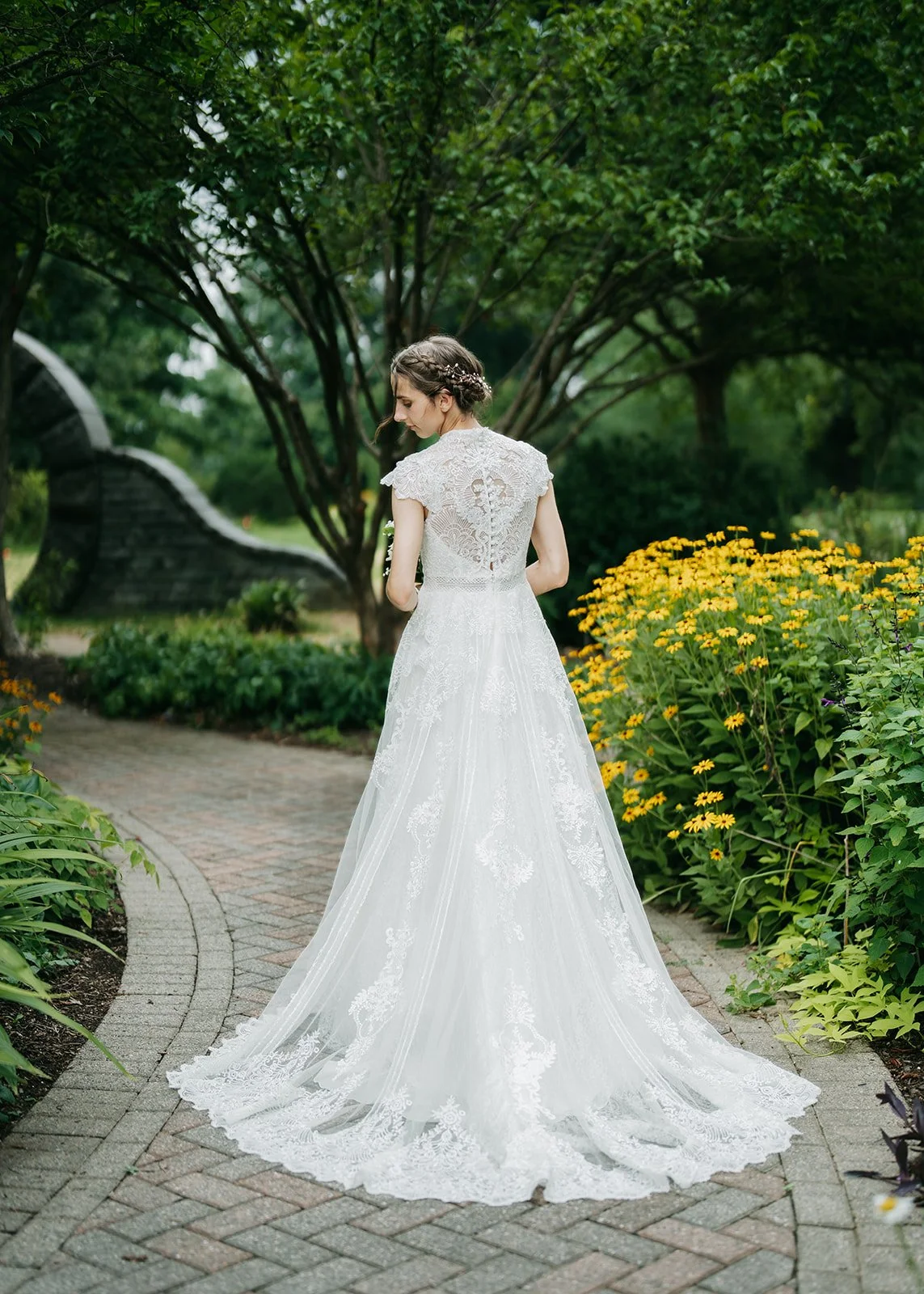 A woman in a white lace wedding gown standing on a brick pathway in a garden, with yellow flowers and green trees around her.